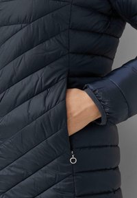 Giacca puffer imbottita blu navy con cuciture diagonali, dotata di tasca laterale con zip e polsini elasticizzati per una vestibilità aderente. Materiale liscio e leggero.