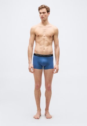 Giovane uomo in piedi a piedi nudi, indossando boxer Polo Ralph Lauren blu con elastico in vita nero, su sfondo grigio chiaro uniforme.