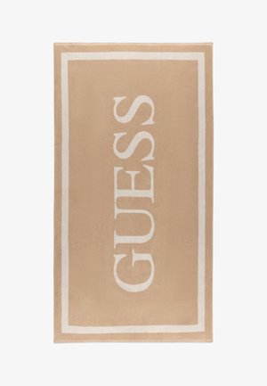 Toalla rectangular beige con borde blanco y el nombre de la marca GUESS impreso verticalmente en grandes letras blancas.