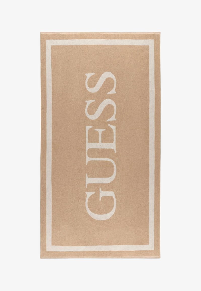 Beige rechthoekige handdoek met witte rand en de merknaam GUESS in grote witte letters verticaal gedrukt.