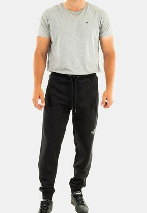 Hombre vestido con camiseta gris de manga corta, pantalones de chándal negros con cordón y zapatillas negras, de pie contra un fondo blanco.