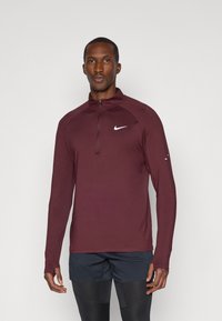 Bărbat îmbrăcat cu un top cu fermoar lung cu mâneci, de culoare vișinie de la Nike, purtând pantaloni scurți negri și colanți, stând în fața unui fundal simplu.