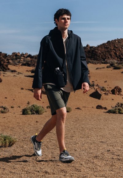 Black jacket, light shirt, olive shorts, and gray sneakers with yellow accents. Casual outdoor attire, walking on sandy terrain.