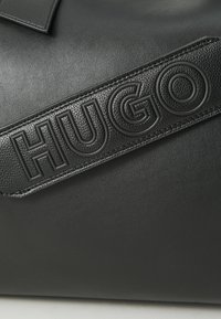 Black leather bag featuring a smooth texture and embossed "HUGO" logo on a wide strap. The overall design is sleek and minimalist.
