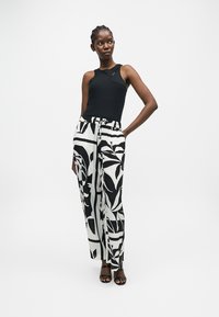 Femme portant un haut noir sans manches et un pantalon large taille haute avec un imprimé botanique noir et blanc audacieux, debout avec une main dans la poche.