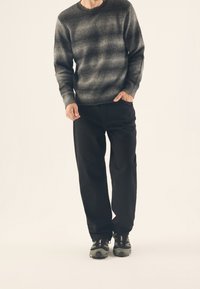 Pull en laine gris à motif dégradé, pantalon noir à coupe décontractée et chaussures de sport noires avec des accents verts. Tenue simple et texturée.