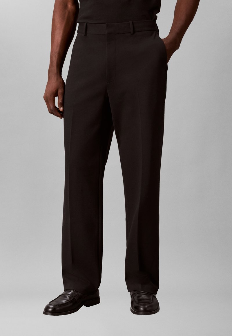 Homme portant un pantalon noir ajusté coupe droite et des mocassins en cuir noir, debout avec une main dans la poche.