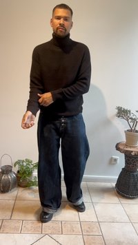 Black knitted turtleneck sweater paired with loose dark denim pants featuring button detail. Model stands on tiled floor with potted plant.