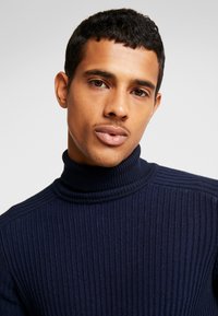 Pull à col roulé côtelé bleu marine avec un motif tricoté texturé et des détails subtils aux épaules. Le tissu semble doux avec une coupe ajustée.