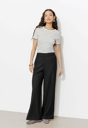 Femme aux longs cheveux foncés portant un t-shirt blanc à manches courtes rayé, un pantalon noir à jambes larges, des ballerines imprimé léopard et de grandes boucles d'oreilles anneau.