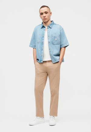 Νέος άνδρας που στέκεται με τα χέρια στις τσέπες, φοράει ανοιχτό μπλε πουκάμισο από τζιν με κοντά μανίκια, λευκή φανέλα, μπεζ παντελόνια και λευκά αθλητικά παπούτσια.