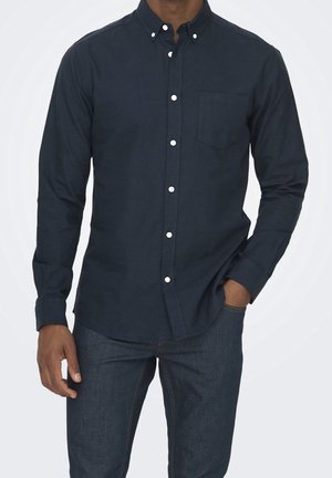 Homme portant une chemise bleu marine foncé à boutons blancs et un jean bleu foncé, une main dans la poche et l'autre détendue à ses côtés.