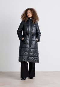 Casaco puffer longo e preto com fecho de zip, cintura com cordão e bolsos laterais; material brilhante e resistente à água com textura acolchoada.