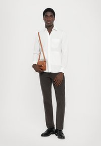 Camicia bianca con bottoni e due tasche sul petto, pantaloni grigio scuro e una borsa a tracolla beige. Scarpe nere completano l'outfit.