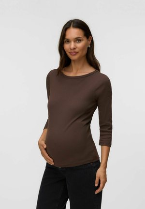Zwangere vrouw die een bruine top met lange mouwen en een zwarte broek draagt, zachtjes haar buik vasthoudend en zacht glimlachend tegen een effen achtergrond.