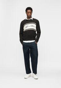 Νέος άνδρας που στέκεται με τα χέρια στις τσέπες, φορώντας μαύρο και λευκό πλεκτό πουλόβερ, λευκό πουκάμισο, σκούρο τζιν και λευκά αθλητικά παπούτσια σε λευκό φόντο.