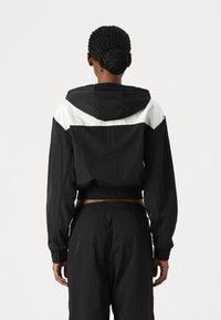 Chaqueta con capucha en blanco y negro con un diseño recortado, hecha de material ligero, que presenta puños elásticos y un distintivo patrón geométrico en la parte posterior.