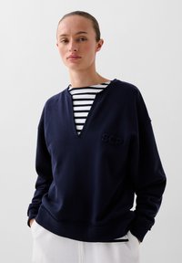 Marineblauwe sweatshirt met een V-hals en gestikte zoom, met het geborduurde "SCP" logo op de voorkant. Draag over een gestreept shirt.