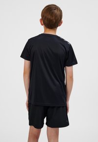 Pojke stående med ryggen mot, iförd en svart kortärmad skjorta och svarta shorts mot en enfärgad ljus bakgrund.