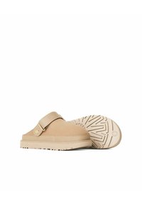 Chaussures à enfiler beiges avec un cuir lisse, une sangle réglable et une semelle en caoutchouc texturée avec un motif de semelle.
