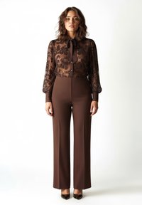 Tuta marrone con una blusa trasparente e decorata con motivi floreali, maniche lunghe e fiocco al collo. I pantaloni sono a gamba larga e di colore marrone solido.