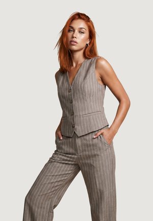Kvinde med rødt hår iført en brun pinstripe ærmeløs vest og matchende vidde bukser, stående med hænderne i lommerne mod en ensfarvet baggrund.
