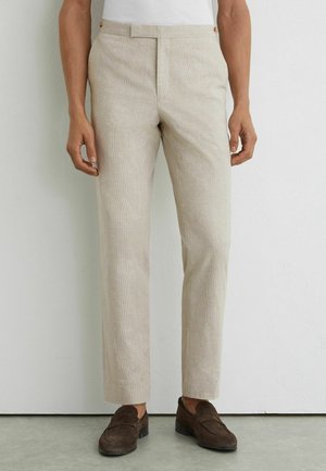 Beige schmal geschnittene Hose mit dezentem Karomuster, kombiniert mit dunkelbraunen Wildleder-Loafern und einem weißen, eingesteckten Hemd.