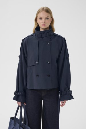 Navy blue cropped jacket with a high collar, double-breasted buttons, wide sleeves, and buckle accents. Model carries a matching tote bag.