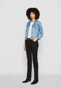 Giacca di jeans blu chiaro con un design aderente, due tasche frontali e bottoni argentati, abbinata a pantaloni neri skinny e stivaletti neri.