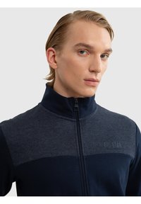 Jonge man met naar achter gestyleerd blond haar draagt een marineblauw jack met rits en het "BIG STAR"-logo op de borst, kijkt iets naar de zijkant.