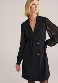 Rochie neagră tip wrap cu mâneci lungi, transparente, curea la talie și accente din nasturi aurii, având un decolteu adânc în V și o croială structurată.