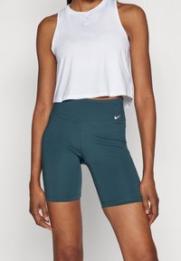 Haut court sans manches blanc avec un col rond. Cuissard de cyclisme taille haute de couleur bleu sarcelle foncé avec des coutures discrètes et le logo Nike sur les deux articles.