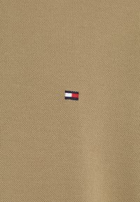 Μπεζ πλεκτό ύφασμα με μικρό κεντημένο λογότυπο με κόκκινες, λευκές και μπλε ρίγες, τοποθετημένο στην αριστερή πλευρά.