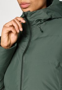 Jachetă verde căptușită cu guler înalt și închidere cu fermoar. Dispune de o textură netedă și detalii de hardware ajustabile în apropierea gulerului.