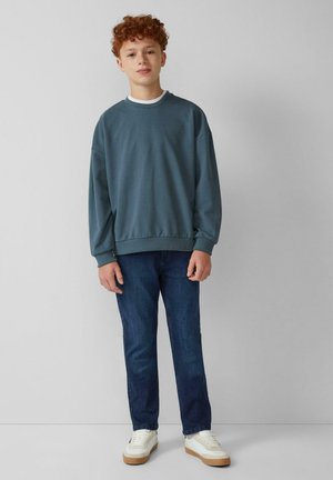 Sweatshirt bleu surdimensionné avec des poignets et un ourlet côtelés, associé à un jean en denim noir et des baskets blanches avec une semelle beige.