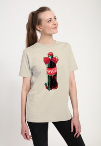 Světle béžové tričko s grafikou lahve Coca-Cola zdobené červenou mašlí a akcenty zimní holly. Krátký rukáv, vyrobené z bavlny.