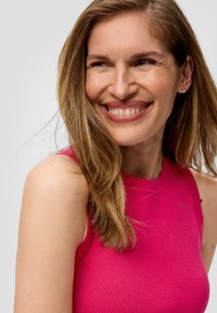 Roze mouwloze top met een ribbelachtige halslijn en een gladde textuur, gedragen door een persoon met lang, golvend bruin haar en minimale oorbellen.