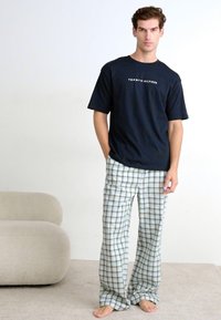 Marineblå t-skjorte med "TOMMY HILFIGER" logo, kombinert med lette rutete pysjamasbukser i blått og krem. Modellen står barbeint med hendene i lommene.