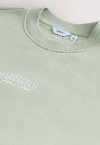 Svijetlozelena majica s rebrastim ovratnikom, s bijelim tekstom "Marrakech" i glatkom, pamučnom teksturom.