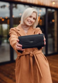 Černá kožená peněženka s hladkým povrchem a zakřiveným víkem, se stříbrným logem jako akcentem. Držena v rukou proti rozostřenému pozadí.