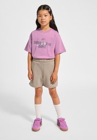 Hellviolettes Baumwoll-T-Shirt mit "Serve the Game" Grafik, kombiniert mit beigen Shorts, weißen Kniestrümpfen und rosa Sportschuhen.