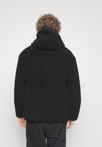 Chaqueta de forro polar negra con capucha, que presenta una superficie texturizada, corte holgado y hombros caídos. La tela texturizada aporta calidez y comodidad.