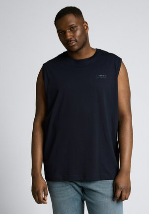 Mężczyzna w czarnej koszulce bez rękawów i jasnoniebieskich dżinsach stoi na tle jednolitego szarego tła.