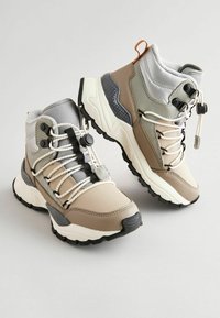 Beige a šedé turistické boty s texturovanou podrážkou, elastickými tkaničkami, polstrovaným límcem a vyztuženými oblastmi pro zvýšenou odolnost.