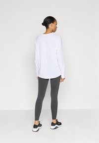 Biela blúzka s dlhým rukávom, zakrivený lem, zadný šev, kombinovaná so sivými legínami a čiernymi teniskami, zobrazená zozadu na jednoduchom pozadí.