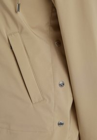 Beige wasserdichte Jacke mit einer Seitentasche, Druckknopfverschlüssen und einem glatten, strukturierten Stoff. Mit silbernem Zubehör akzentuiert.
