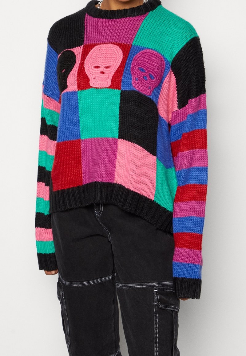 Maglione multicolore lavorato a maglia con un motivo a blocchi, caratterizzato da tre design di teschi. Collo a girocollo e polsini a coste; colori vivaci includono nero, verde, rosa e viola.