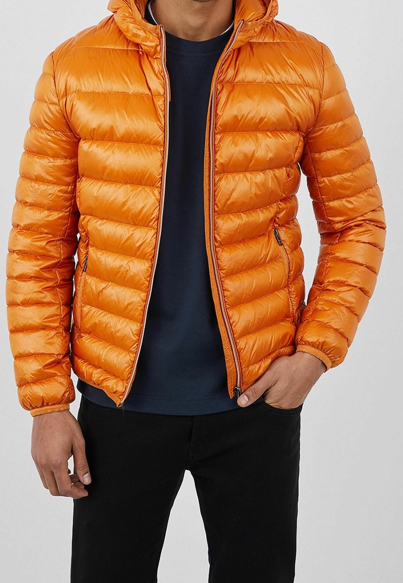 Homme portant une veste matelassée orange vif sur une chemise bleu marine foncé avec un pantalon noir, une main dans la poche, fond neutre.