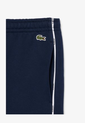 Námornícke modré tepláky vyrobené z jemnej tkaniny, s malým zeleným logom krokodíla, bielymi bočnými pruhmi s brandingom "LACOSTE" a elastickým pásom.