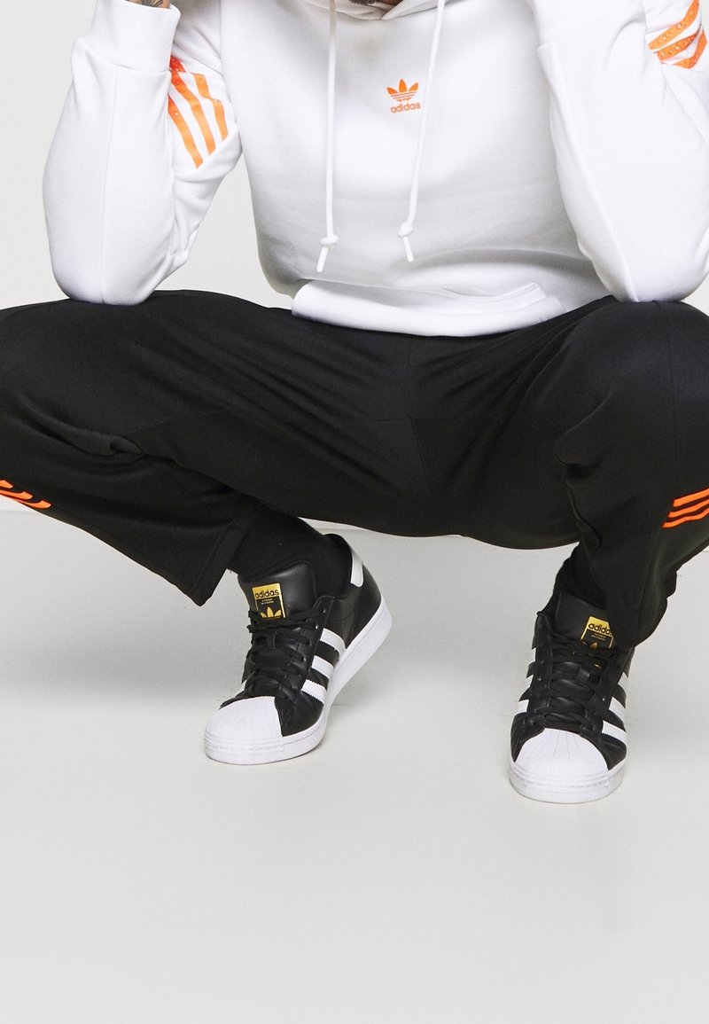 Vit hoodie med orange detaljer, svarta träningsbyxor och svarta Adidas Superstar sneakers med vita och gyllene detaljer.